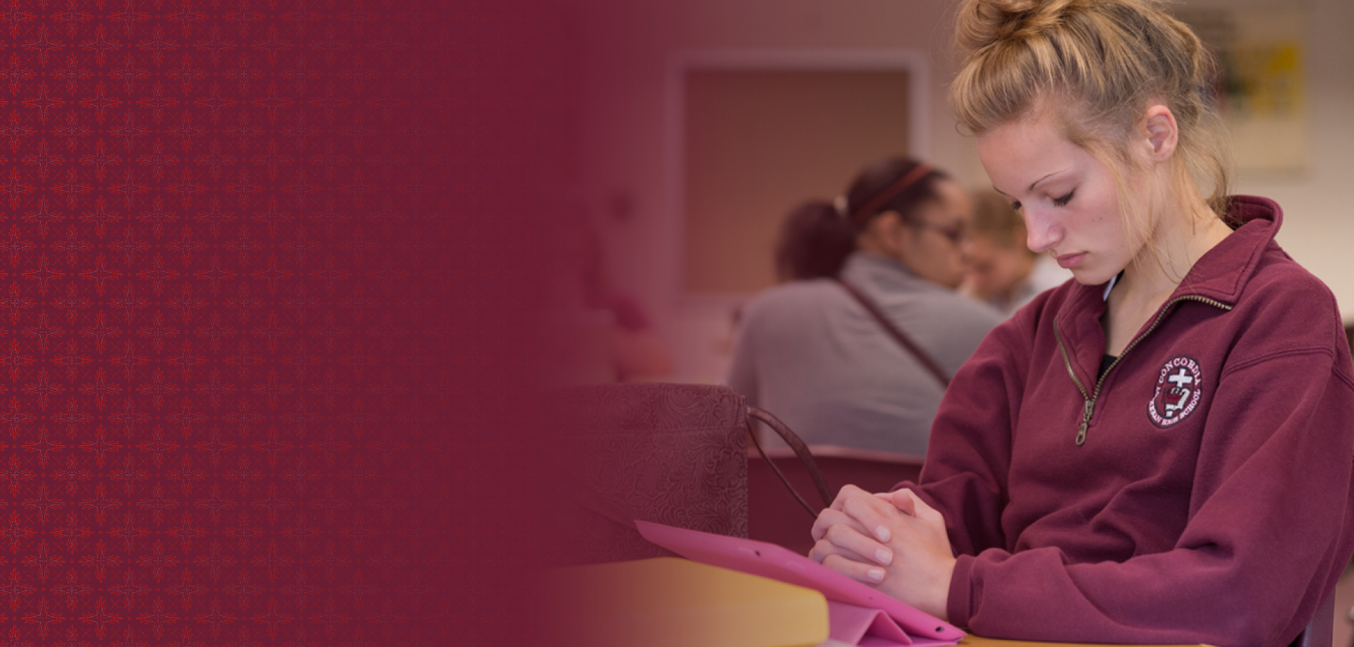 Student in a maroon hoodie studying at a table, looking down at a pink tablet or folder in front of her.