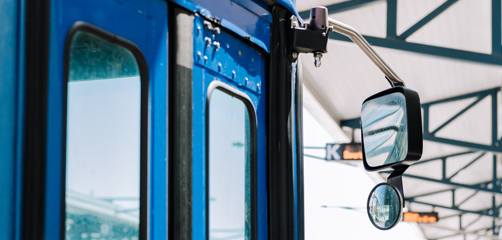 Close-up of a blue bus side with a large side mirror reflecting the station roof and windows.