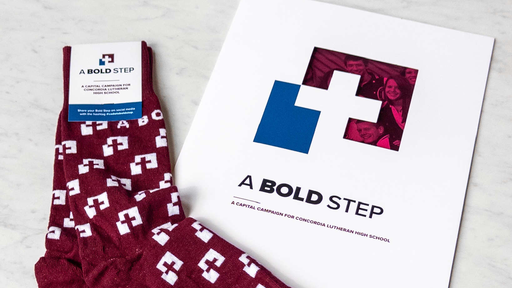 Branding materials for 'A Bold Step': white sheet with blue-and-white cross logo and magenta photo area, beside a maroon sock with white cross pattern.