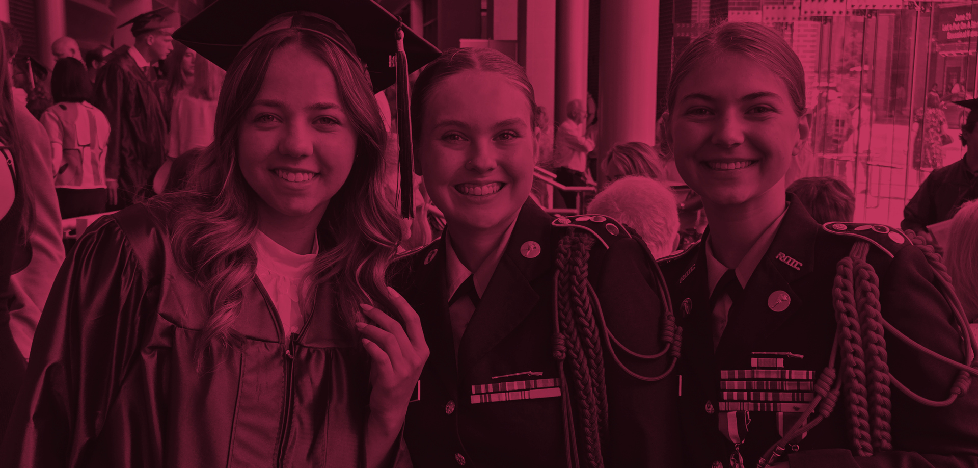 Three smiling graduates posing for a photo: a woman in a cap and gown on the left, and two women in formal uniforms with medals and cords on the right.