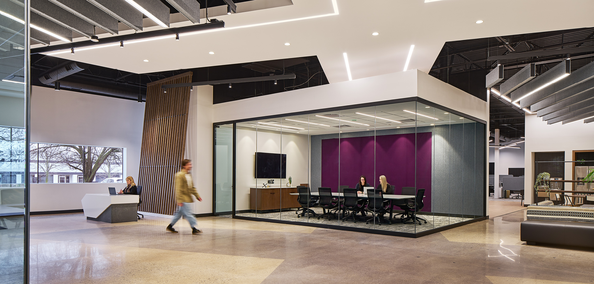 Open-plan corporate lobby featuring a glass-walled conference room with a purple accent wall and people seated around a long table inside the room.