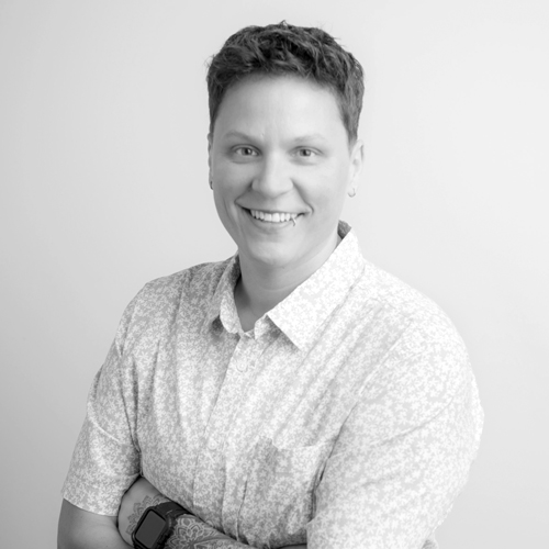 Portrait of a smiling person with short hair, wearing a light patterned shirt and a smartwatch on their left wrist, arms crossed.