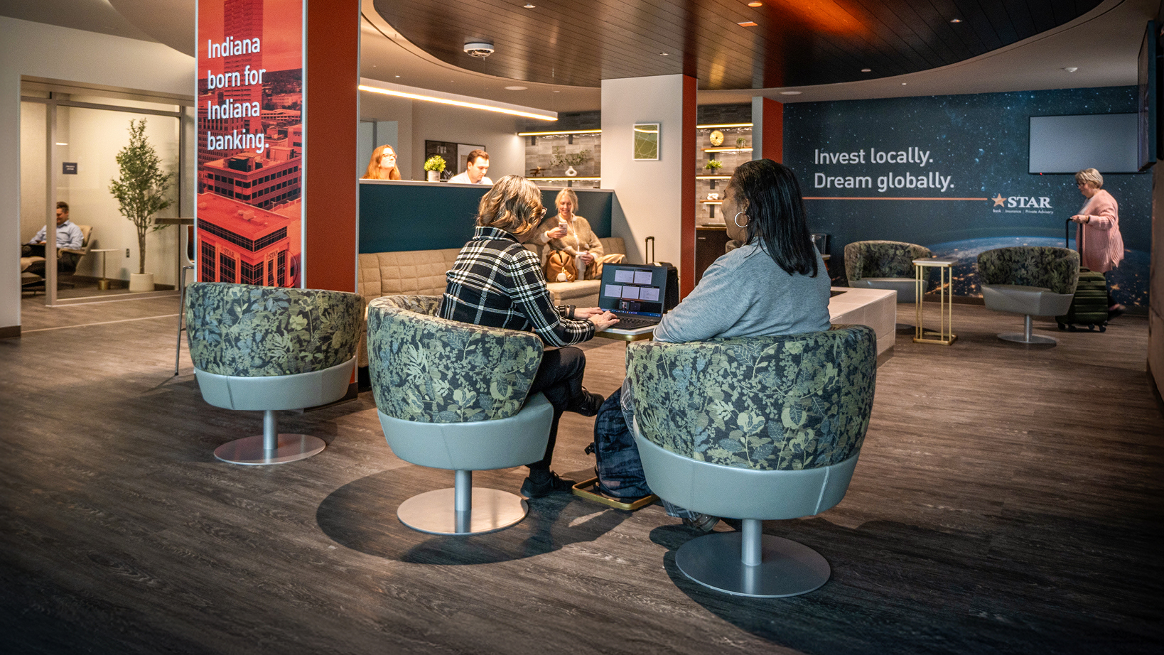 Two women sit on upholstered circular chairs in a bank lounge, one using a laptop as they talk.