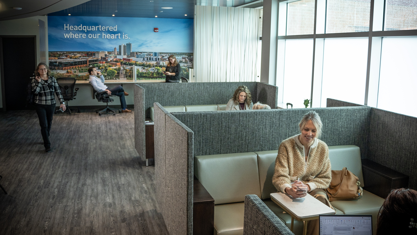 Modern office lounge with gray fabric partitions; a woman in a beige sweater sits at a small table using her phone, bag beside her, near large windows.