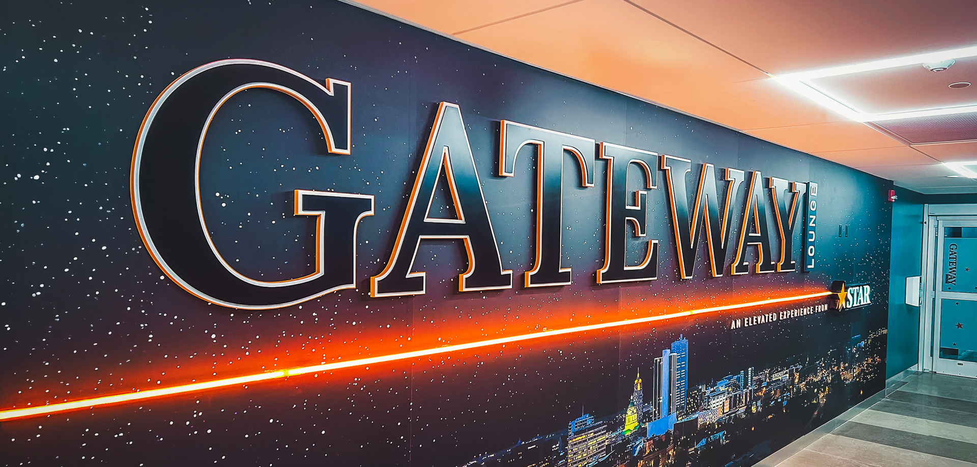 Large 3D 'GATEWAY' sign on a space-themed wall with a city skyline at the bottom and an orange neon strip running along the base.