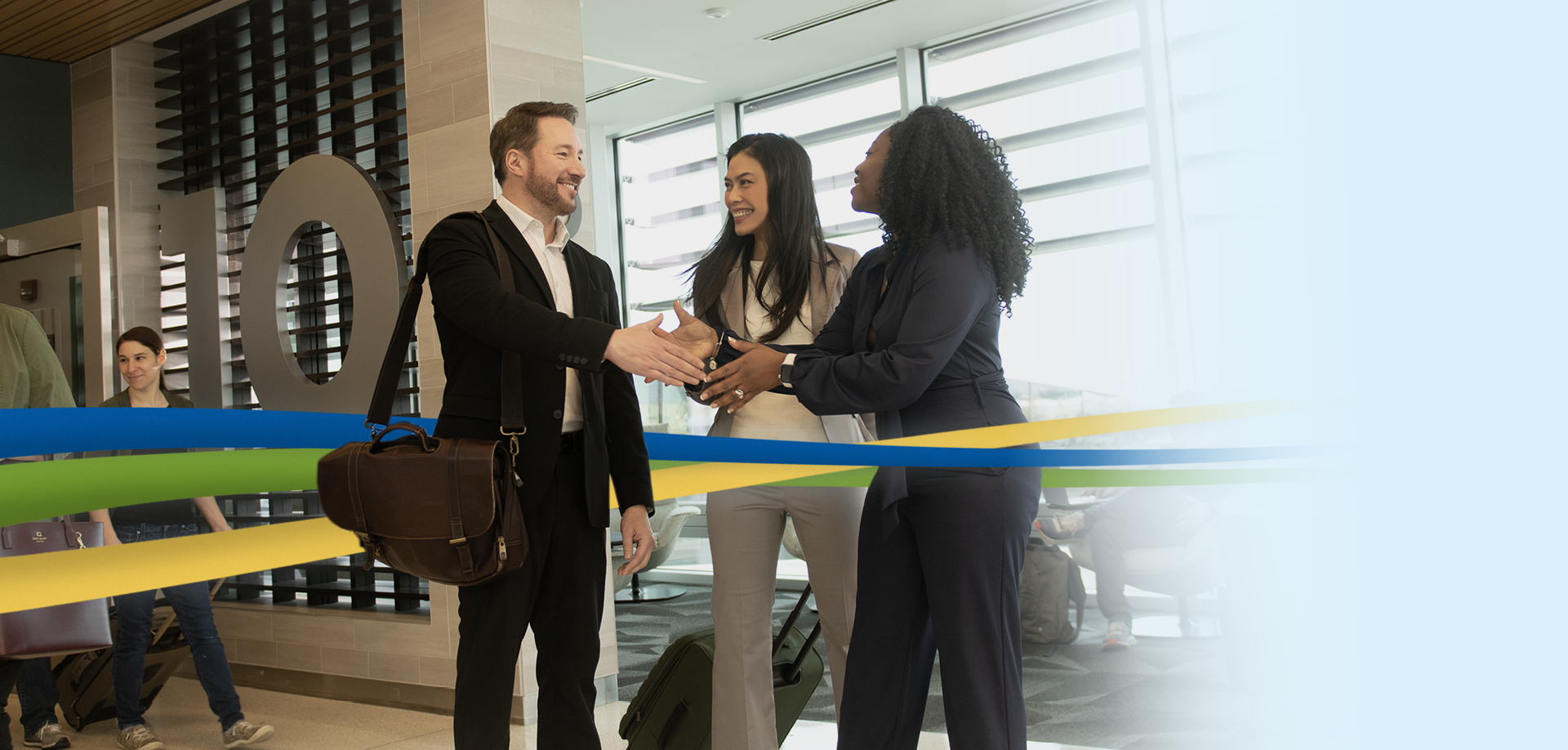 Man in a suit shakes hands with a woman in a dark blazer as a colleague smiles nearby in a modern lobby with colored ribbons and luggage nearby.
