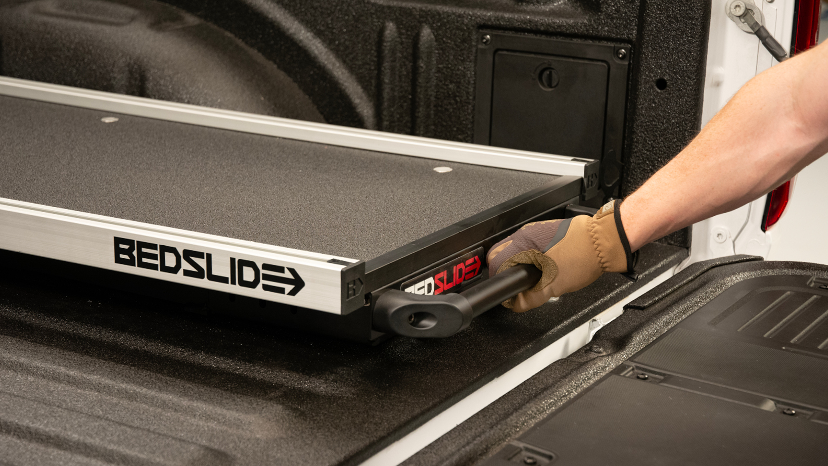 Gloved hand pulls a BedSlide drawer out of a pickup bed, extending it from the truck bed rails.