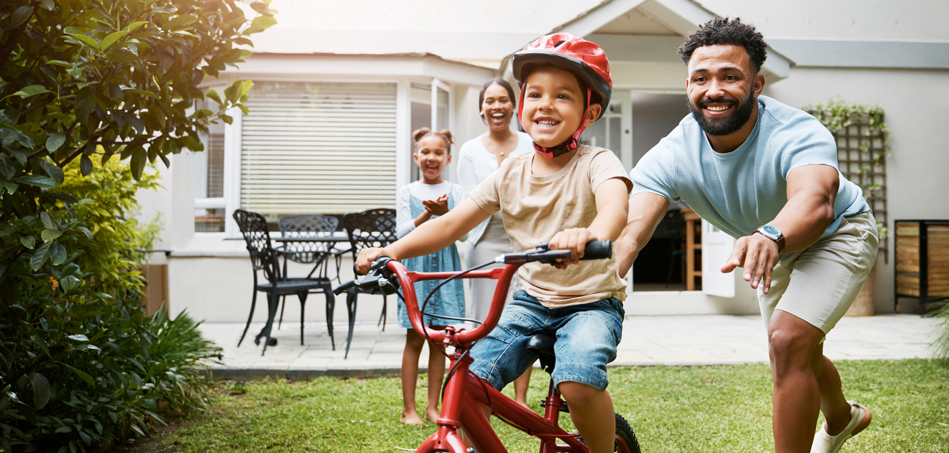 Dad helps a smiling boy ride a red bike in the yard as family members cheer in the background.
