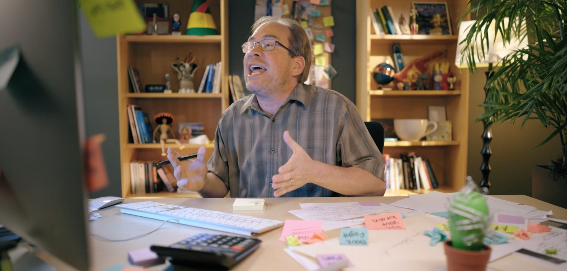A man with glasses sits at a cluttered desk, gesturing as he speaks, with a computer monitor and colorful sticky notes nearby. shelves and a plant are in the background.