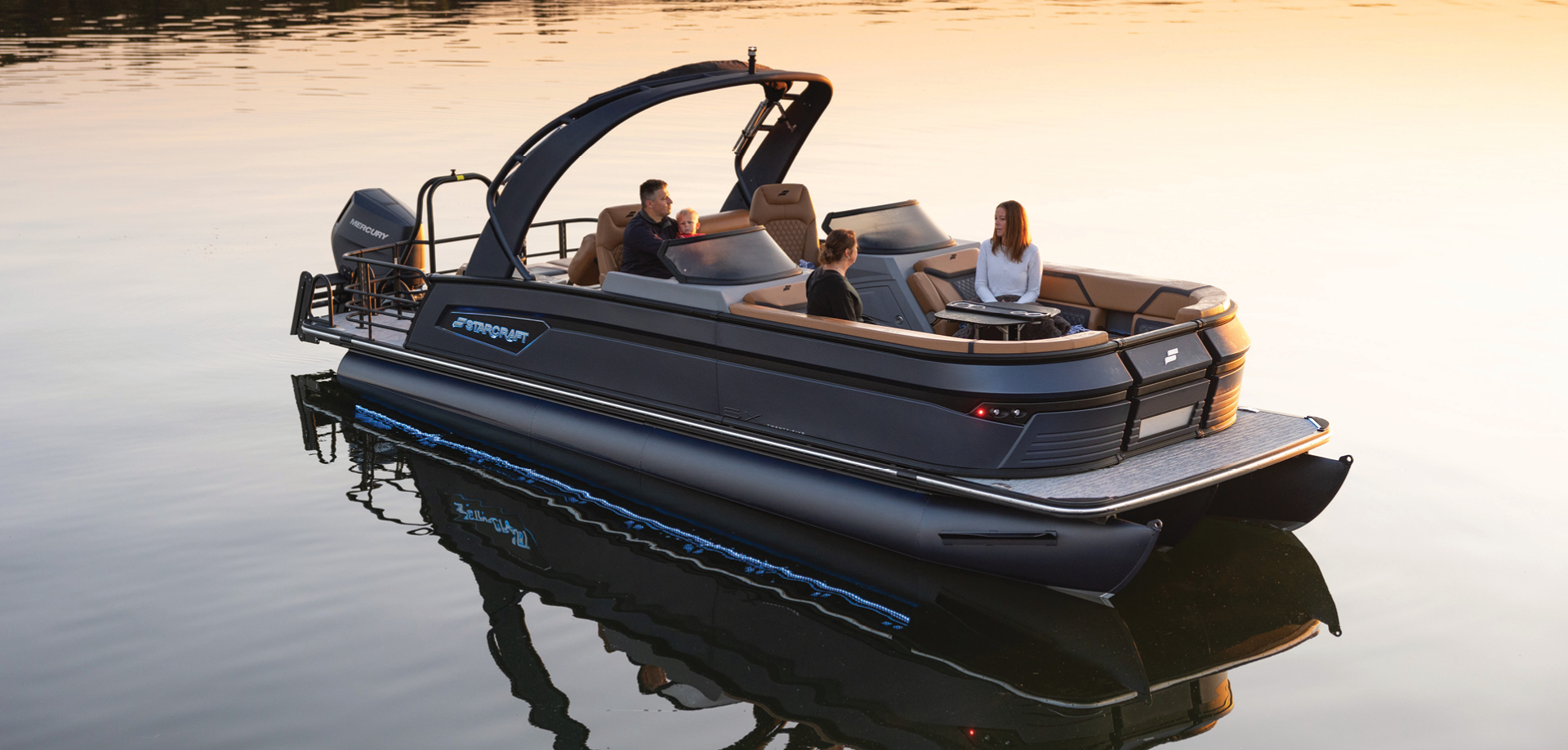 Dark gray Starcraft pontoon boat with tan seating carrying four people on a calm lake at sunset