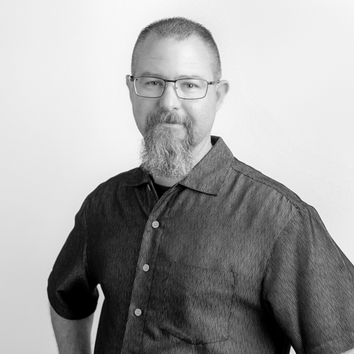 Portrait of a man with short hair, goatee and glasses, wearing a dark button-down shirt and looking at the camera.