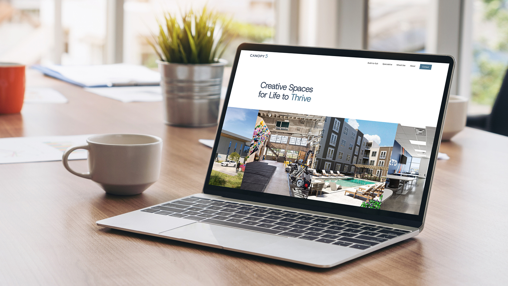 Laptop on a wooden desk showing a website hero with the slogan 'Creative Spaces for Life to Thrive' and a collage of modern interior spaces outside the top navigation bar.