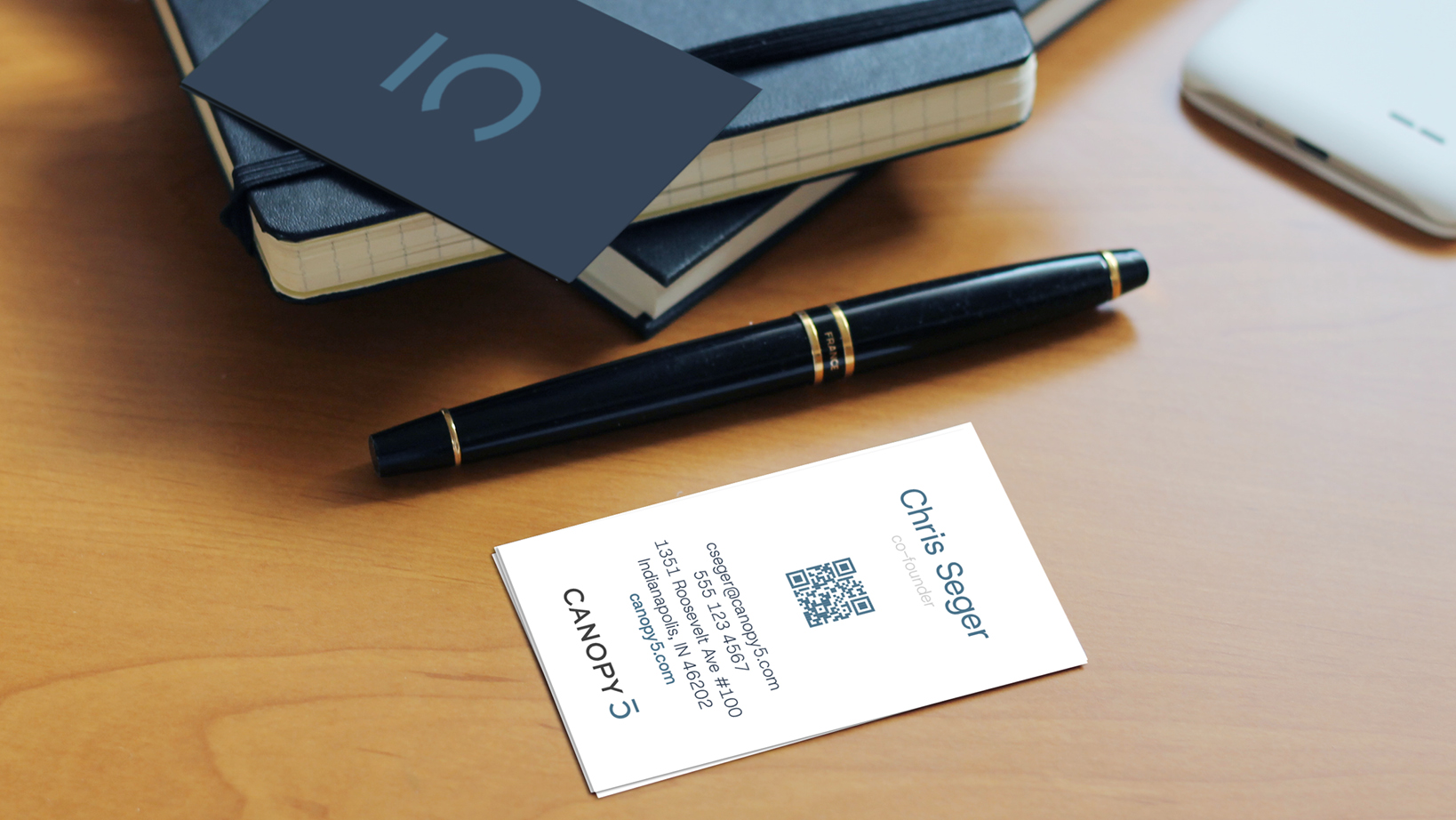 Close-up of a desk with a black pen, stacked notebooks, and a white business card showing the name 'Chris Seger' and a QR code.