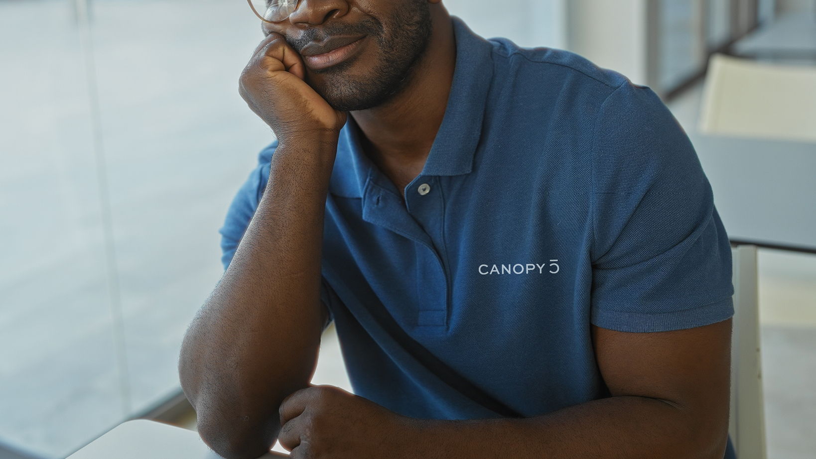 Close-up of a man in a blue polo resting his chin on his hand, CANOPY logo on his chest.