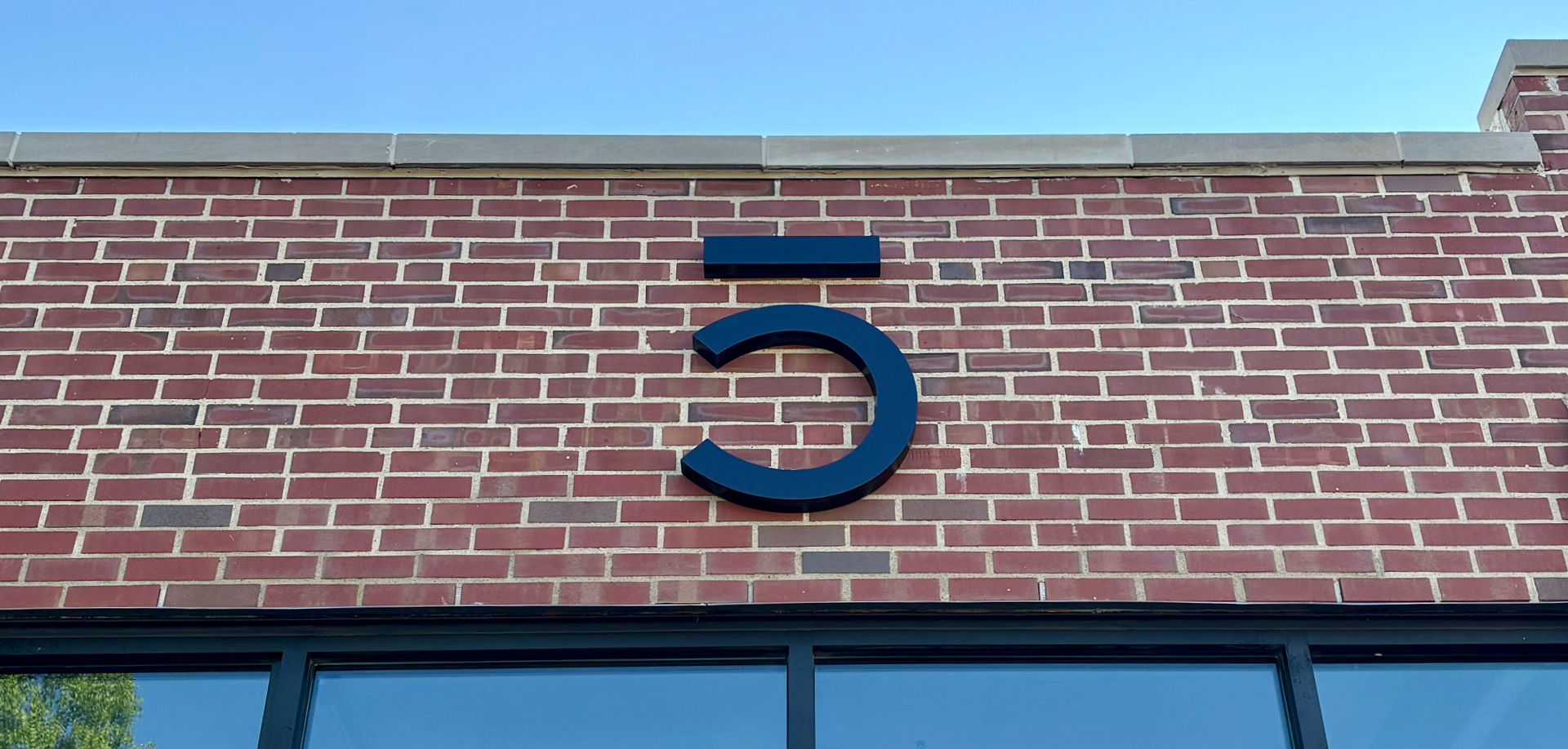 Large black stylized number 5 mounted on a red brick storefront wall against a clear blue sky.