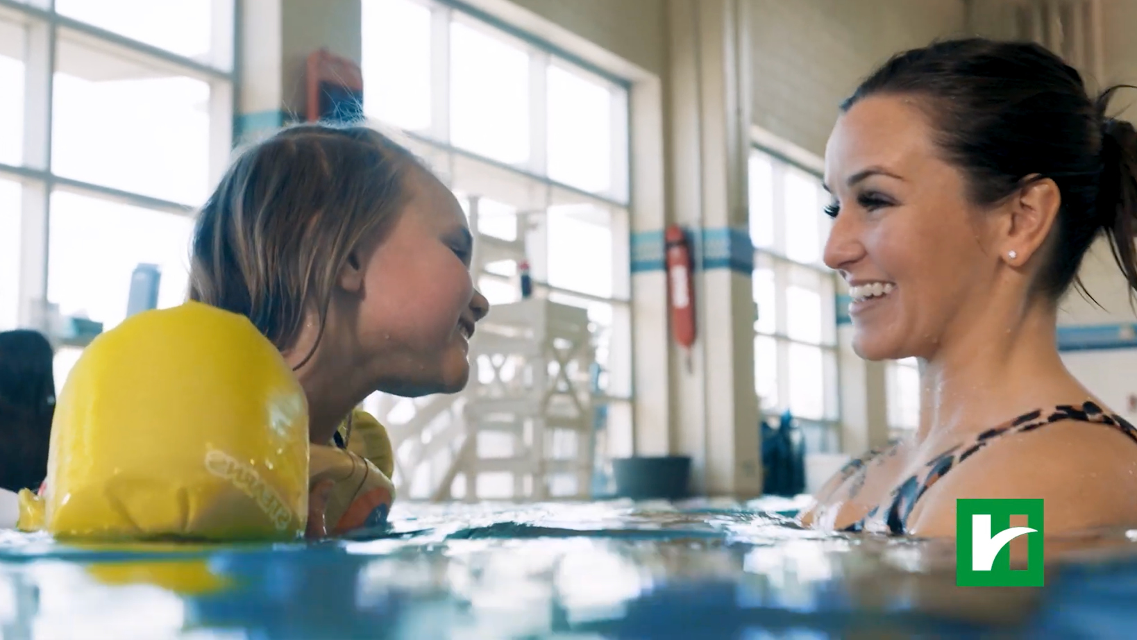 Child in a yellow life vest smiles at a female swim instructor in an indoor pool during a lesson.