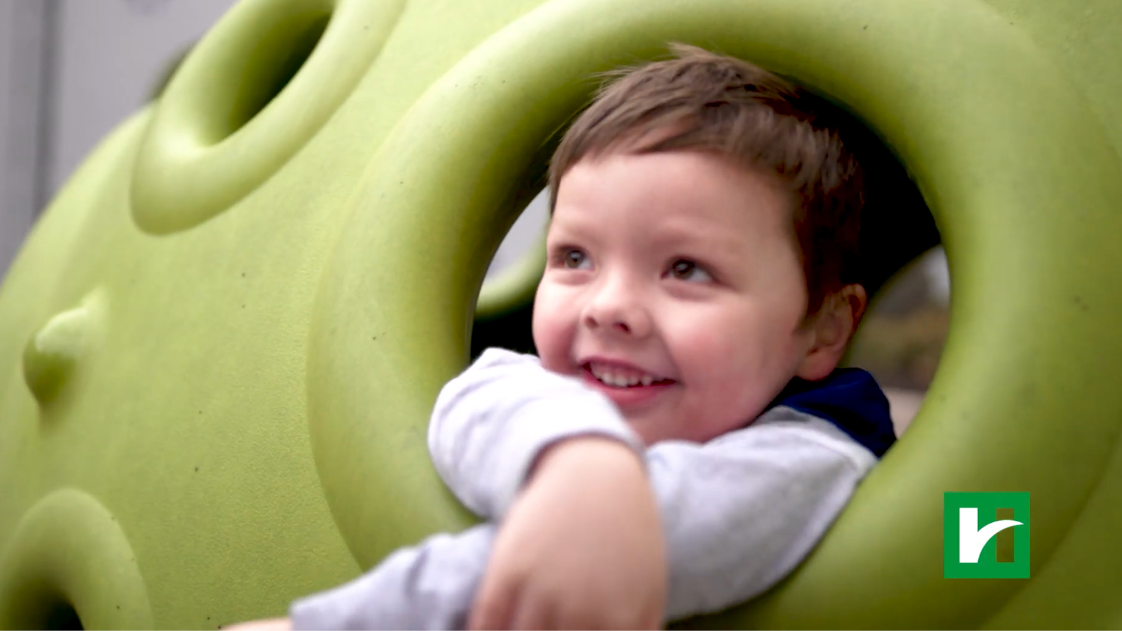 Smiling child peeks through a lime-green spiral playground structure, looking upward.