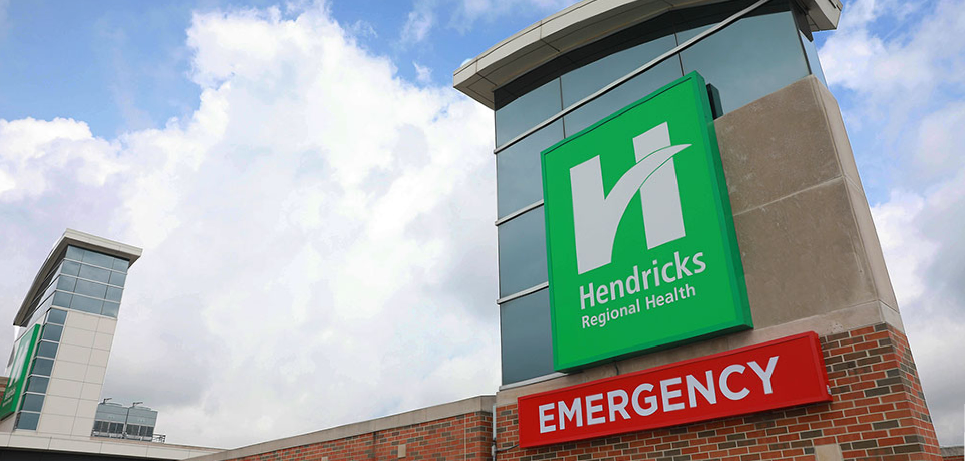 Exterior view of a hospital entrance with a bright green Hendricks Regional Health sign and a red EMERGENCY sign on a brick wall.