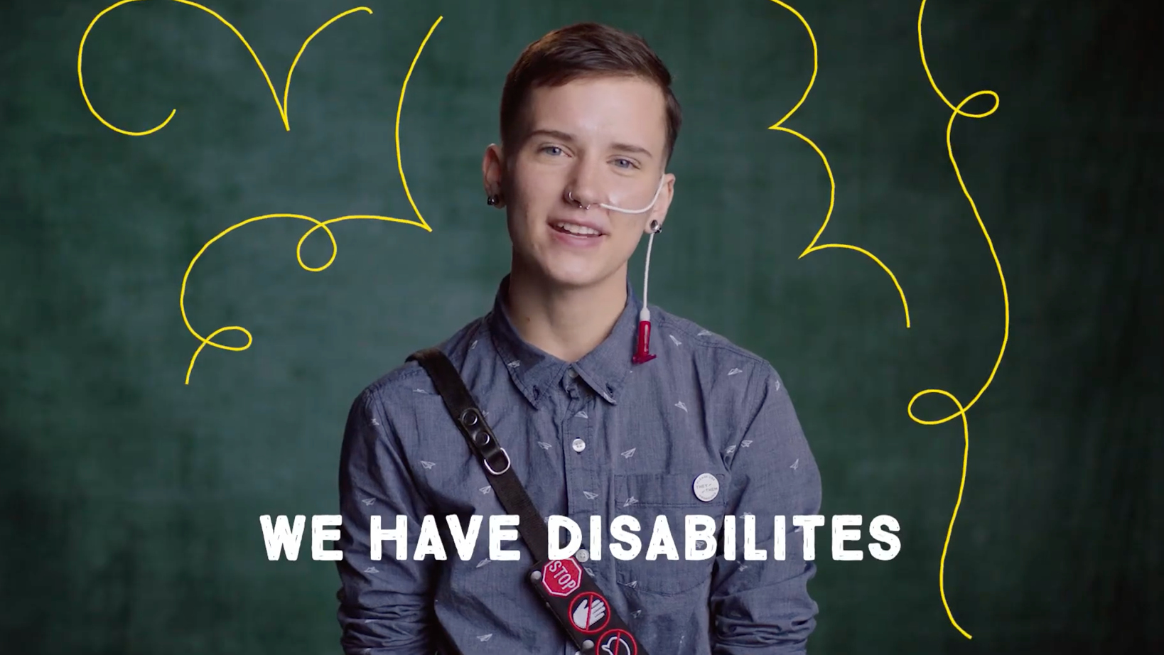 Person with a tracheostomy speaks to the camera in a blue shirt; chalkboard backdrop with bright yellow doodles and the caption 'WE HAVE DISABILITIES'.
