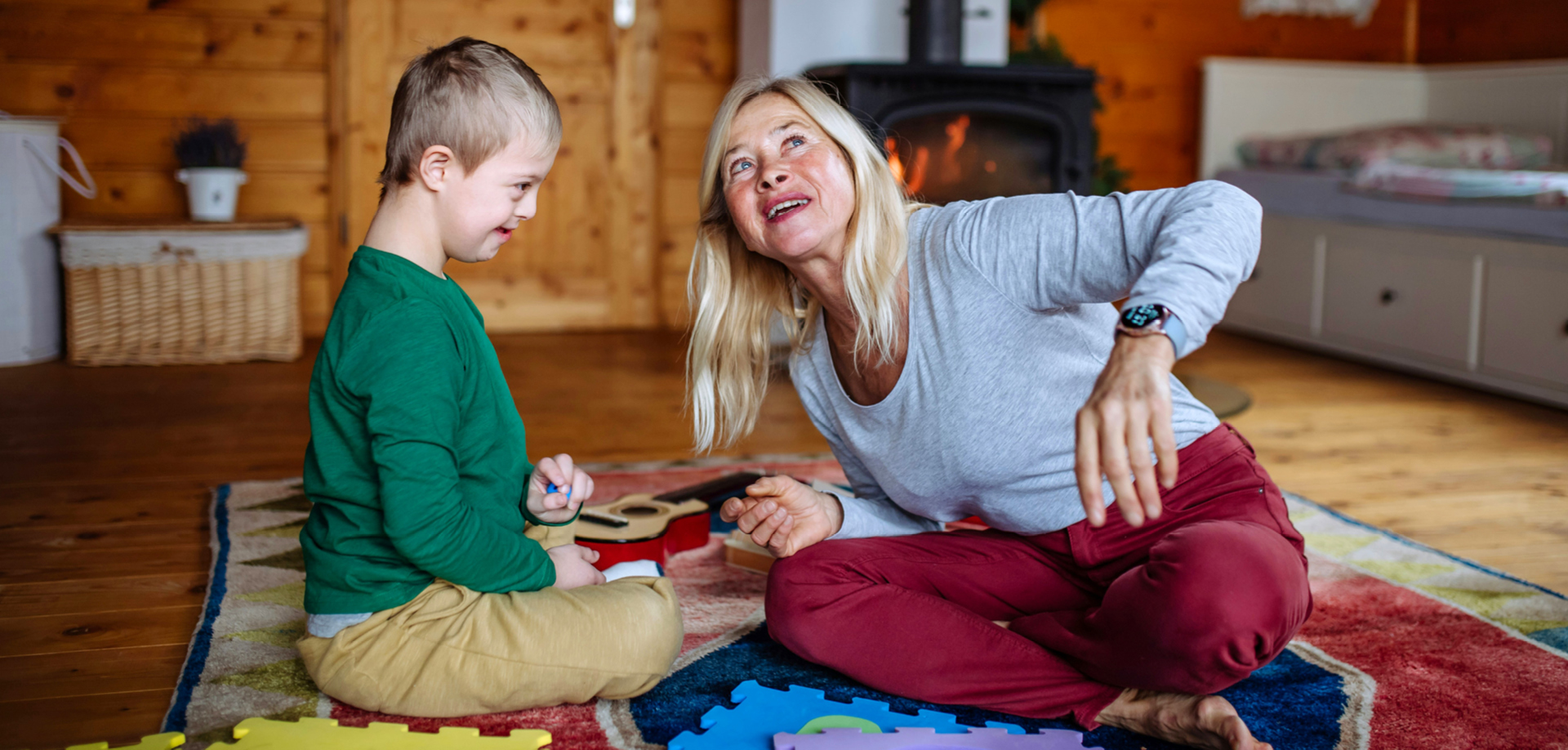 Woman and young boy sit on a colorful rug, playing with puzzle pieces in a cozy wooden living room by a fireplace.