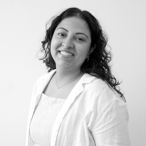 Smiling woman with dark, wavy hair wearing a white blouse and necklace, standing against a light background.