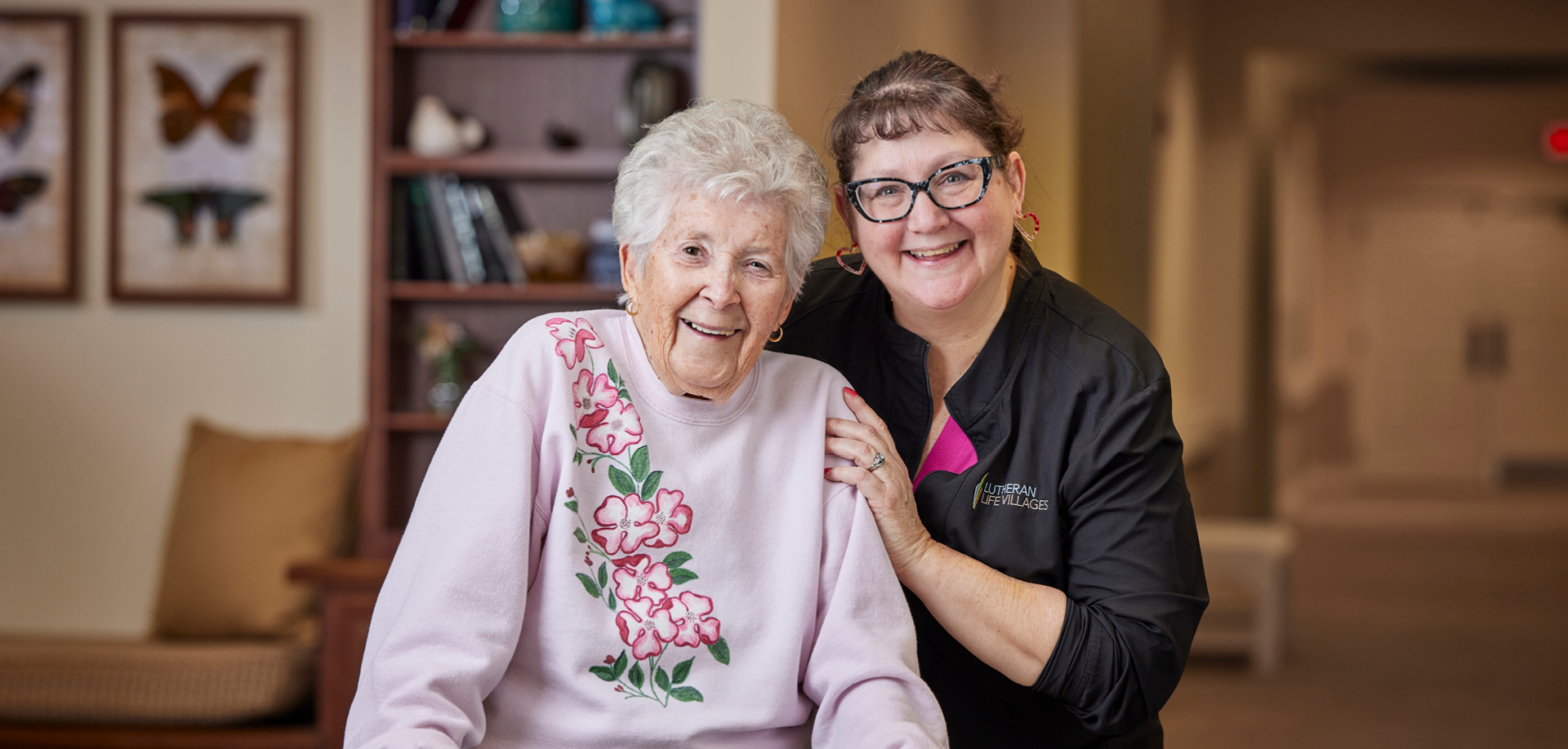 Smiling elderly woman in a pink floral sweater posing with a caregiver in a cozy living room setting.