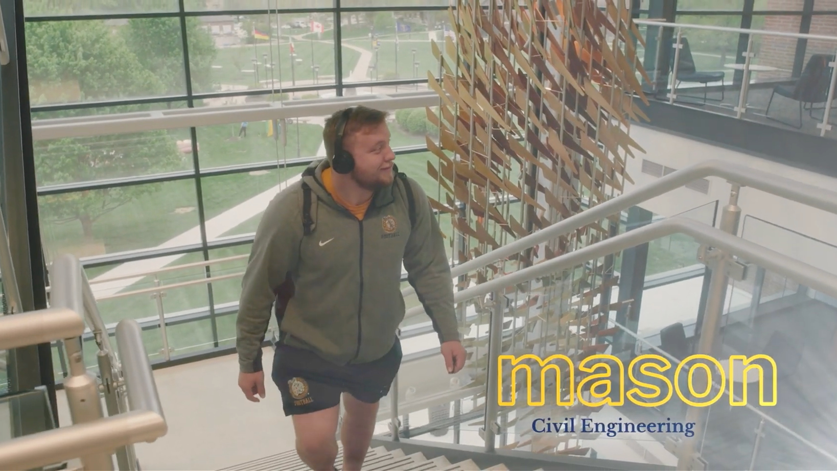 Man wearing a green hoodie with a logo and headphones, ascending a glass staircase inside a modern building; campus visible outside the large windows; overlay shows 'mason Civil Engineering'