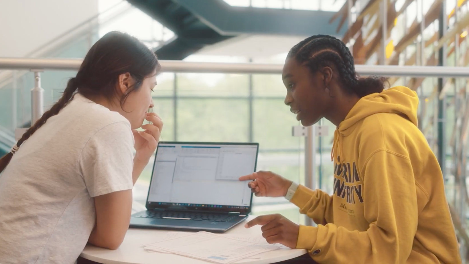 Two students discuss a laptop screen at a round table, papers spread out in a bright indoor study space.