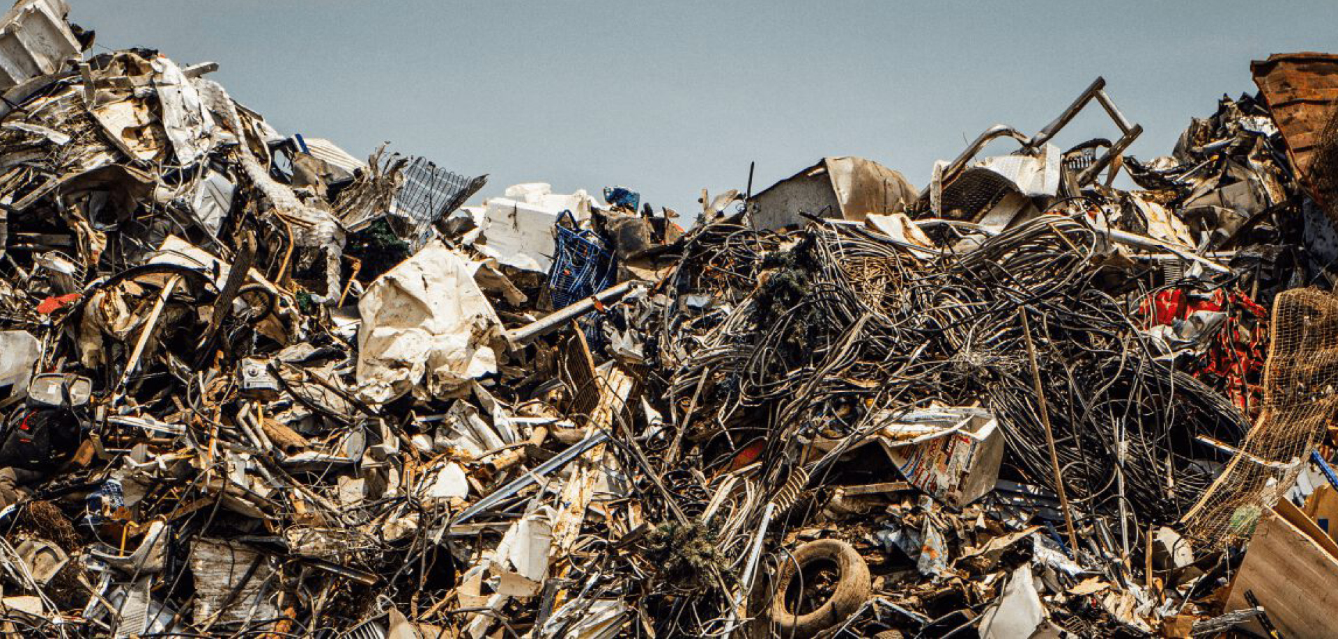 Large mound of tangled scrap metal, wires, and debris in a scrapyard against a pale blue sky