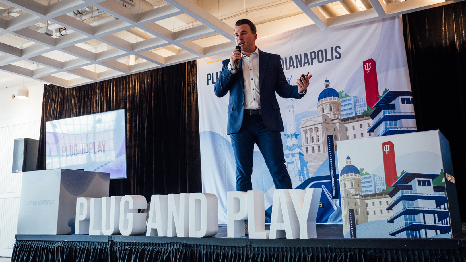 Male speaker in a navy suit speaks into a handheld microphone on stage, with large 'PLUG AND PLAY' letters at the front and an Indianapolis-themed backdrop behind him.