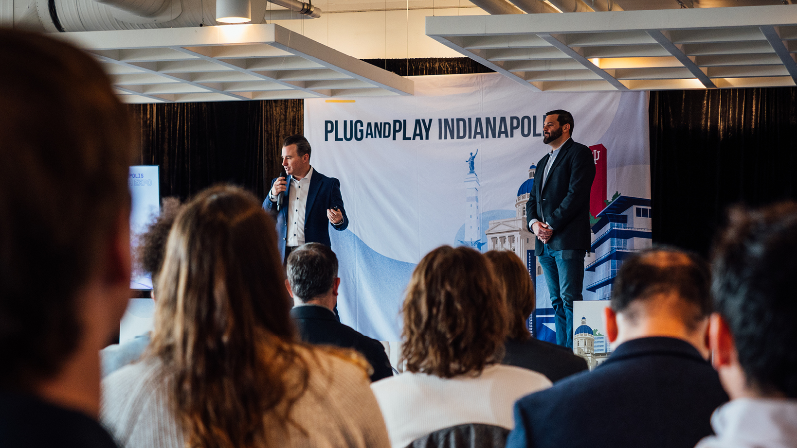 Two men on a stage at Plug and Play Indianapolis; one speaks into a microphone while the other stands beside him, with an audience in the foreground and a cityscape banner behind them.