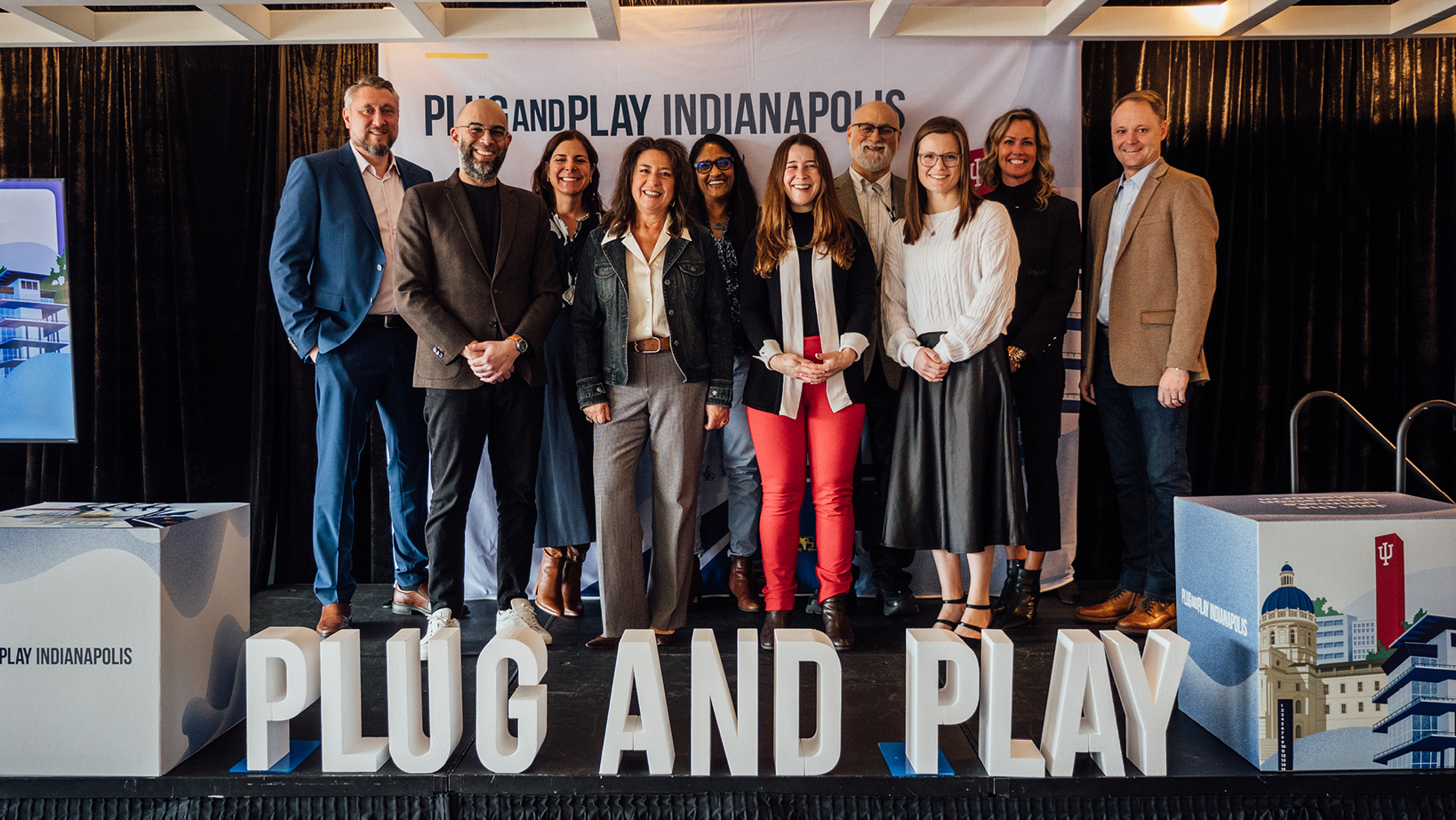 Group of professionals posing on a stage at a Plug and Play Indianapolis event, with the event banner in the background.