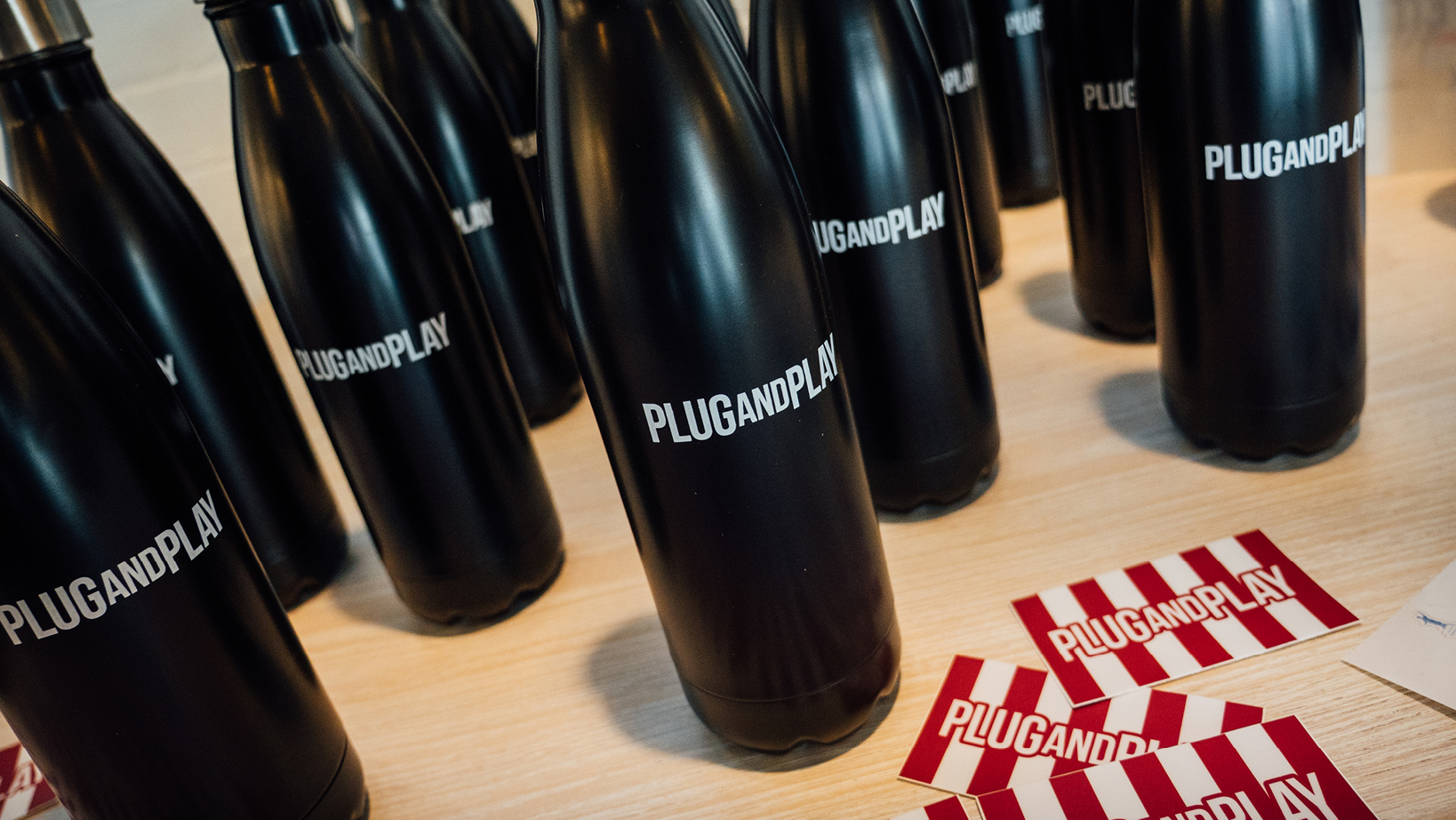 Row of black bottles with 'PLUGANDPLAY' branding on a light wooden table, with red-and-white striped brand cards nearby.