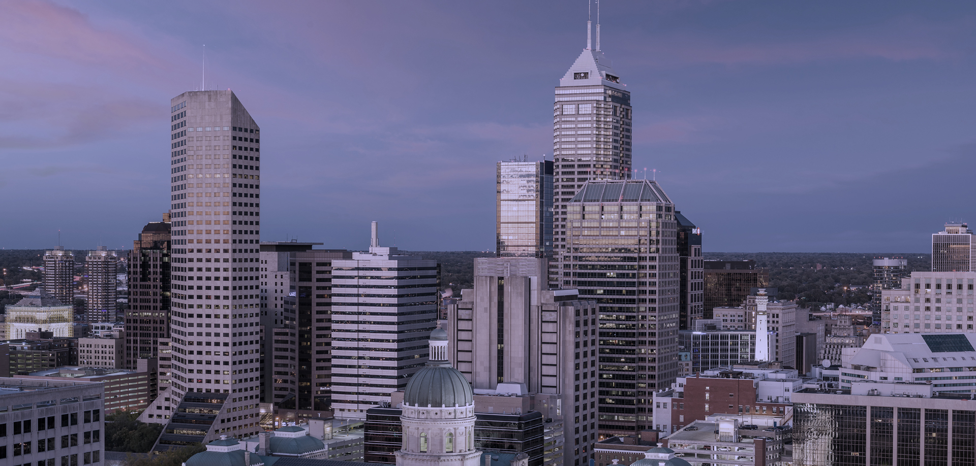 City skyline at dusk with tall modern skyscrapers and a domed historic building in the foreground.
